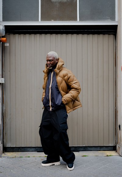 Chaqueta acolchada marrón con capucha sobre una chaqueta azul marino con cremallera, combinada con pantalones cargo negros y zapatillas de deporte gruesas en negro y blanco. Fondo urbano.