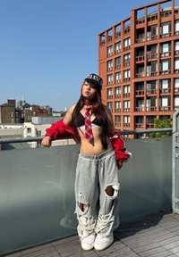 Röd bomberjacka, svart crop top, rutigt slips. Grå slitna sweatpants med stora hål, vita sneakers. Urban miljö med byggnader.