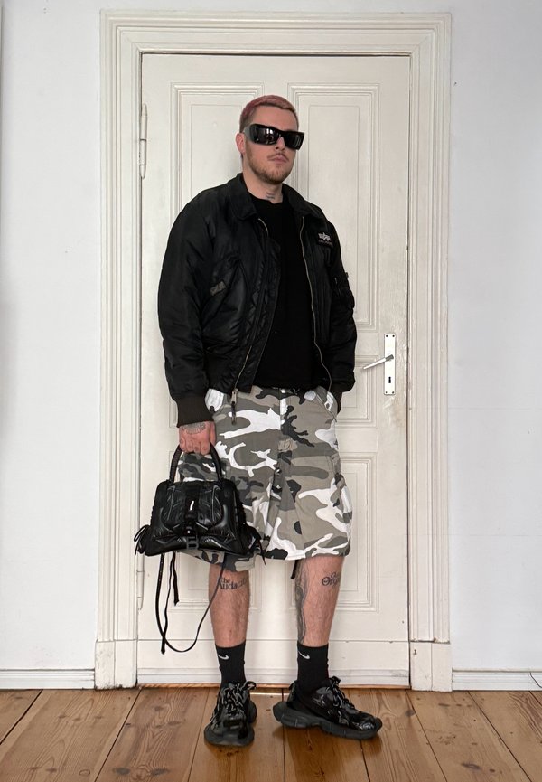 Homme aux cheveux courts teints portant une veste noire, une chemise noire, un short camouflage, des baskets noires et des lunettes de soleil, tenant un sac à main noir à l'intérieur près d'une porte blanche.