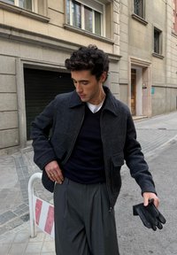 Giacca da uomo in lana grigio scuro con colletto, tasche frontali e cerniera; abbinata a un maglione nero e pantaloni grigi plissettati. Guanti neri tenuti in mano.