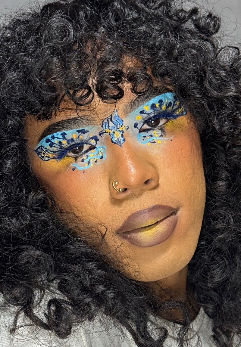 Nahaufnahme einer Person mit lockigem, dunklem Haar, detailliertem, blauer und gelber, schmetterlingsinspirierter Augen-Make-up, das sich über die Augenbrauen und die Nase erstreckt, und matten Lippen.