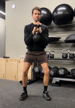 Hombre levantando una pesa rusa con ambas manos a la altura del pecho en un gimnasio, rodeado de balones de ejercicio, pesas rusas y bolsas con peso.