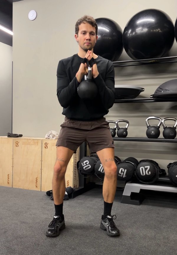 Homme soulevant une kettlebell avec les deux mains au niveau de la poitrine dans une salle de sport, entouré de ballons d'exercice, de kettlebells et de sacs lestés.