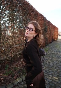 Jeune femme portant des lunettes et une tenue noire, posant avec un geste de baiser sur un chemin pavé à côté d'une haute haie de feuilles brunes en plein jour.