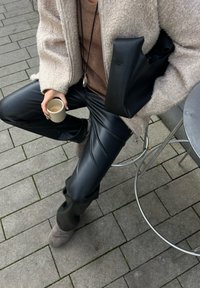 Pantalones de cuero sintético negros, abrigo beige texturizado, suéter marrón, sosteniendo una taza beige. El fondo presenta un pavimento de piedra gris y una mesa de metal.