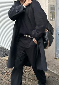 Dlhý čierny kabát cez tmavý outfit, doplnený čiernym opaskom. Osoba drží čiernu koženú tašku, pričom okolie tvorí dlažobná kocka.