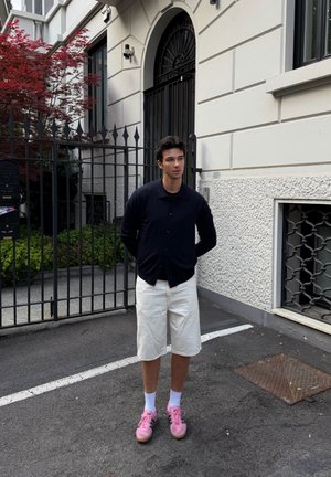 Joven está en la calle frente a la entrada con puerta cerrada de un edificio, lleva camisa negra, pantalones cortos blancos, calcetines blancos y zapatillas rosas.