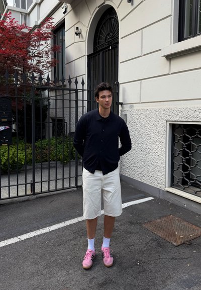Joven está en la calle frente a la entrada con puerta cerrada de un edificio, lleva camisa negra, pantalones cortos blancos, calcetines blancos y zapatillas rosas.