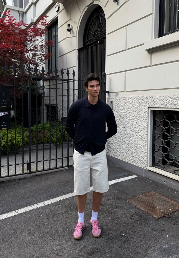 Mladi muškarac stoji na ulici ispred ulaza u zgradu s kapijom, nosi crnu košulju, bijele kratke hlače, bijele čarape i ružičaste tenisice.