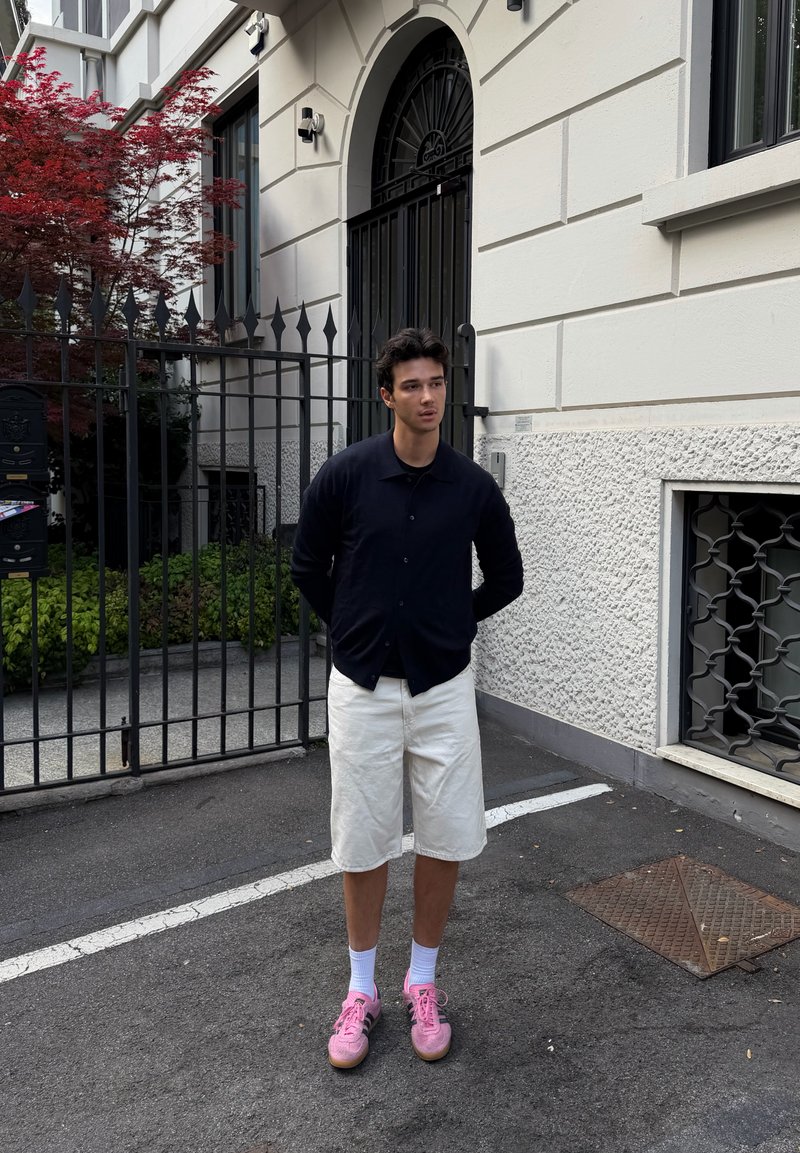 Jeune homme se tient dans la rue devant l'entrée grillagée d'un bâtiment, portant une chemise noire, un short blanc, des chaussettes blanches et des baskets roses.