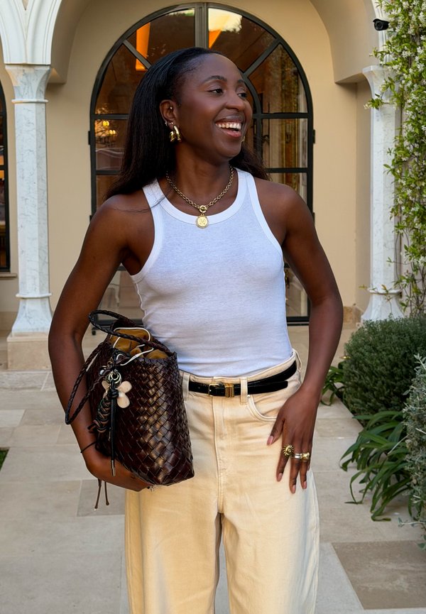 Lächelnde Frau in weißem Tanktop und cremefarbenen Hosen hält eine geflochtene dunkelbraune Handtasche und steht draußen in der Nähe eines gewölbten Fensters mit grünem Bewuchs.