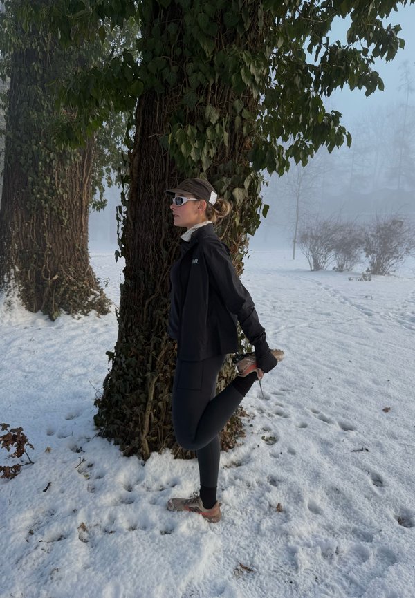 Femme en tenue de course d'hiver étire une jambe derrière elle à côté d'un arbre dans un parc enneigé et brumeux.