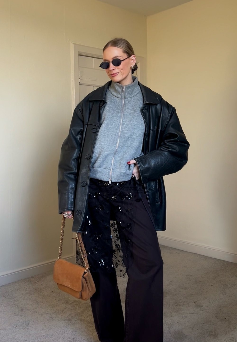 Femme portant une veste en cuir noire oversize, un pull gris à fermeture éclair, un pantalon noir orné, des lunettes de soleil noires, tenant un sac à main beige à l'intérieur.