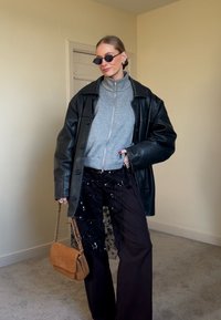 Femme portant une veste en cuir noire oversize, un pull gris à fermeture éclair, un pantalon noir orné, des lunettes de soleil noires, tenant un sac à main beige à l'intérieur.