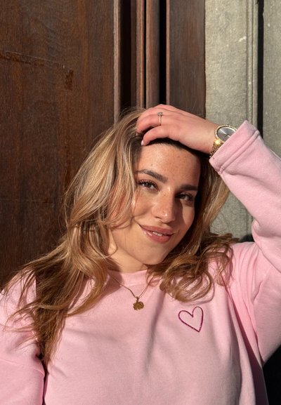 Sudadera rosa claro con bordado de corazón, combinada con un collar dorado. El cabello está estilizado en ondas sueltas, frente a un fondo de madera.