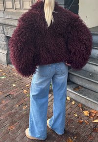 Pessoa com cabelo loiro longo, vestindo uma jaqueta felpuda castanha escura, calças largas azuis claras e sapatos de plataforma bege, numa calçada de tijolos.