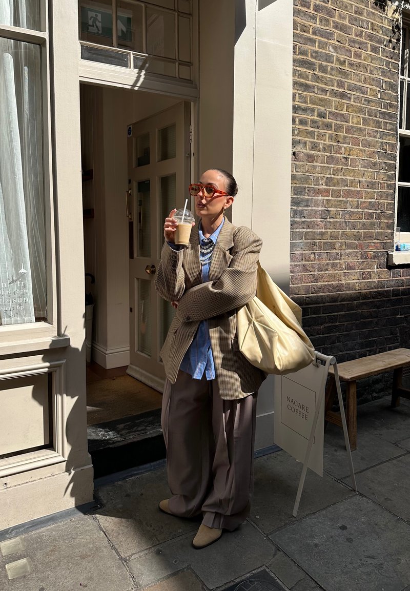 Eine Person in einem übergroßen beigen karierten Blazer, blauen Hemd und grauen Hosen hält ein Getränk. Sie trägt rote Brille und eine cremefarbene Tote-Bag.