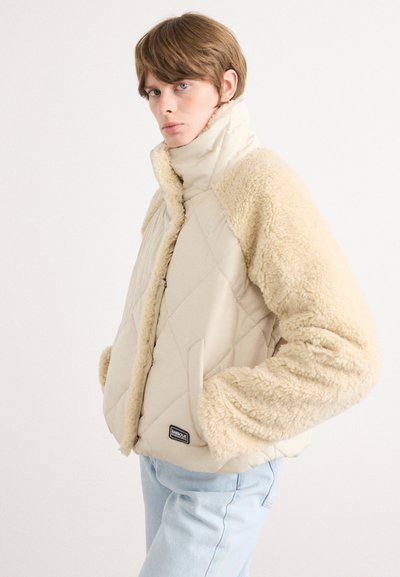 Chaqueta acolchada beige con mangas de borrego crema, cuello alto y cierre de cremallera. Presenta una pequeña etiqueta negra en la parte frontal. Corte casual.