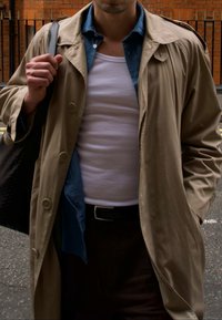 Tan trenchcoat over en blå skjorte og en hvit tanktop. Svart veske i hånden. Knappet jakke med bred krage og glatt stoff.