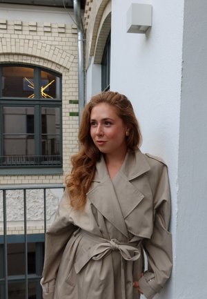 Trench coat beige con un cinturón y solapas anchas, de mangas largas y con una textura suave. El fondo presenta una pared blanca y una ventana.