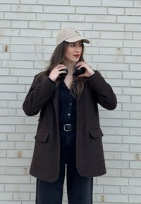 Jeune femme ajustant un casque noir, portant une casquette beige, un manteau texturé foncé, une ceinture noire et un pull en maille bleu marine, debout devant un mur de briques clair.