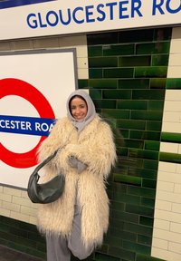 Personne souriante en manteau beige en fausse fourrure avec capuche, tenant un sac noir, debout à la station de métro Gloucester Road à Londres.