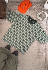 Groen gestreept t-shirt met blauwe en oranje lijnen, voorzien van een donkere kraag en het "VANS"-logo. Gecombineerd met een oranje pet en witte koptelefoon.