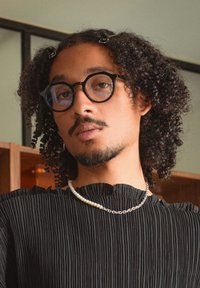 Top negro con pliegues, cuello redondeado. Gafas negras, collar de perlas y cadenas. Cabello rizado adornado con clips para el pelo, fondo neutro.