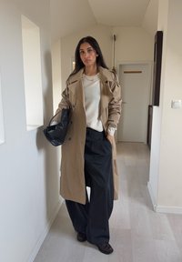 Trench-coat beige porté sur un pull crème et un pantalon large noir, tenant un sac noir. Textures lisses, design croisé remarquable.