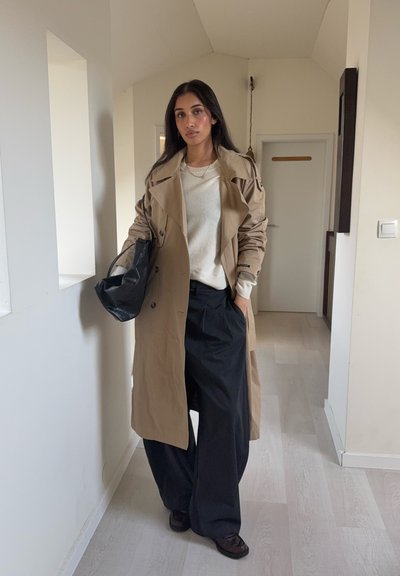 Gabardina beige sobre un suéter crema y pantalones anchos negros, sujetando un bolso negro. Texturas suaves, diseño notable de doble botonadura.