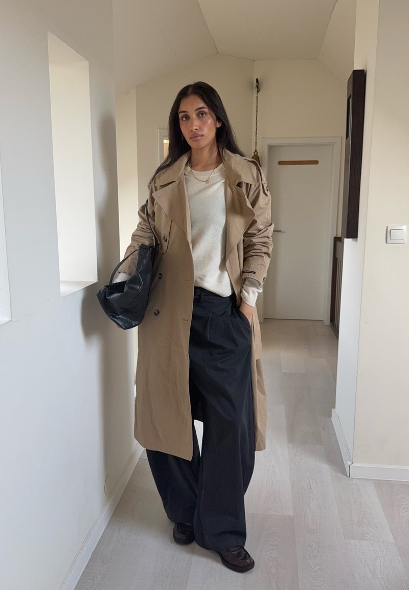 Trench-coat beige porté sur un pull crème et un pantalon large noir, tenant un sac noir. Textures lisses, design croisé remarquable.