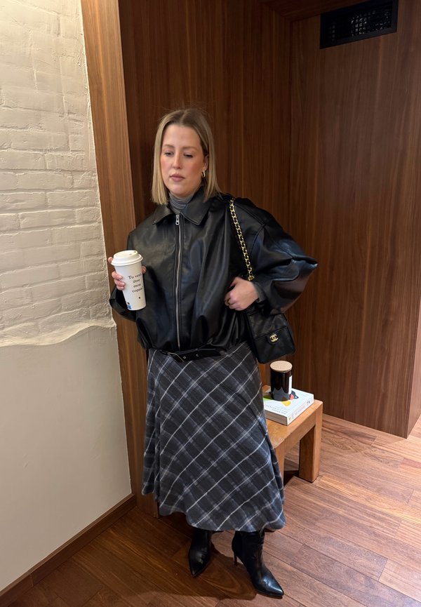 Donna con giacca di pelle nera, gonna a quadri grigia e stivali neri, che tiene una tazza di caffè e una borsa nera in una stanza con pareti rivestite in legno.