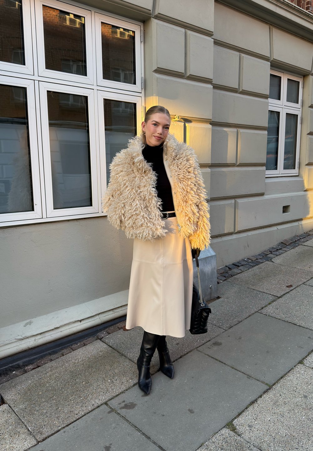 Femme debout sur le trottoir portant une veste en fausse fourrure beige, un col roulé noir, une jupe midi crème, des bottes noires et tenant un sac à main noir.