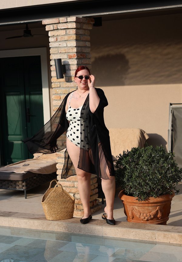 Femme portant un maillot de bain blanc à pois et une tunique noire transparente, debout près de la piscine, ajustant ses lunettes de soleil, avec un sac tressé et une plante en pot à proximité.