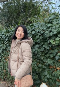 Femme souriante en plein air portant une doudoune beige et tenant un sac à main beige, debout devant un feuillage dense de lierre vert.
