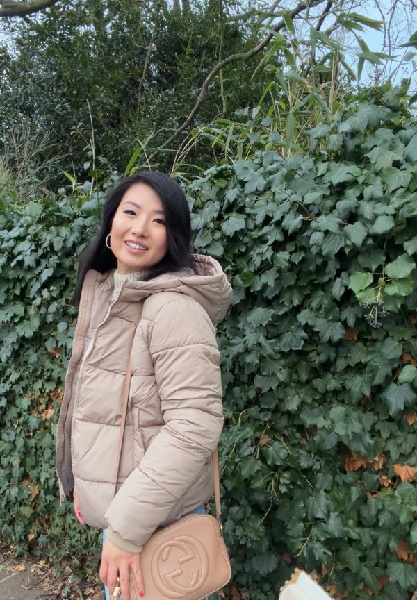 Femme souriante en plein air portant une doudoune beige et tenant un sac à main beige, debout devant un feuillage dense de lierre vert.