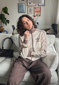 Casaco de pista Adidas em creme com detalhes e riscas marrons, combinado com calças largas marrons. Os acessórios incluem pulseiras e brincos de ouro.