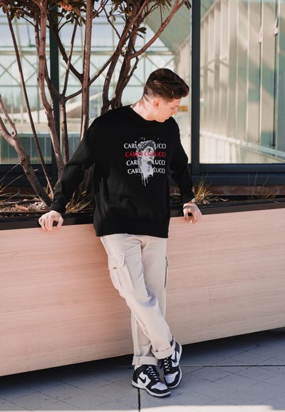 Sudadera negra con estampado gráfico, pantalones cargo beige y zapatillas negras y blancas. El escenario presenta macetas y paneles de vidrio.