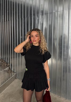 Conjunto de dos piezas negro con frunces, compuesto por una parte superior ajustada y pantalones cortos acampanados, realzado con un broche verde, combinado con medias texturizadas y un bolso rojo.