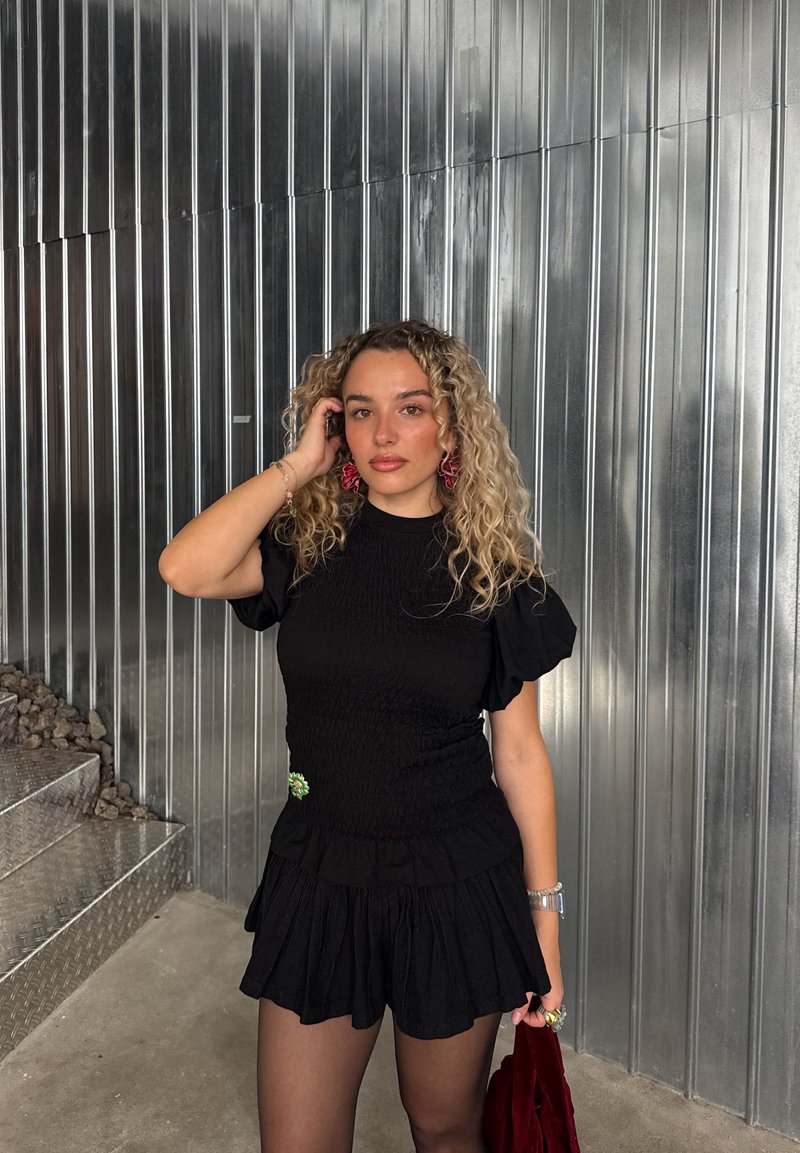 Conjunto de dos piezas negro con frunces, compuesto por una parte superior ajustada y pantalones cortos acampanados, realzado con un broche verde, combinado con medias texturizadas y un bolso rojo.