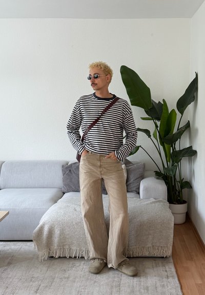 Persona con cabello rubio rizado que lleva gafas de sol, camisa de rayas, pantalones beige y bolso bandolera, está de pie en una sala de estar con sofá y una planta grande.