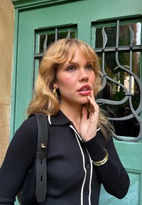 Nero maglietta a costine con bottoni e accenti bianchi, orecchini a cerchio dorati e una borsa nera. Porta verde con lavori in ferro decorativi sullo sfondo.