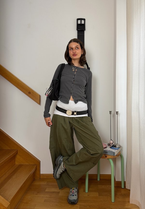 Mujer apoyada contra una pared con un cinturón negro extragrande, lleva una camiseta gris, una camisa blanca, pantalones verdes, zapatillas plateadas y lleva una bolsa negra en el hombro.