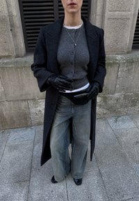 Femme portant un long manteau sombre, un pull gris boutonné, un jean à jambes larges, des gants noirs, un collier et un sac ceinture noir, debout sur un pavement en pierre.
