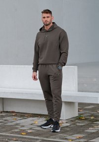 Sweat à capuche vert olive foncé avec cordons ajustables et joggeurs assortis. Tissu texturé, associé à des chaussures de sport noires avec des accents orange.