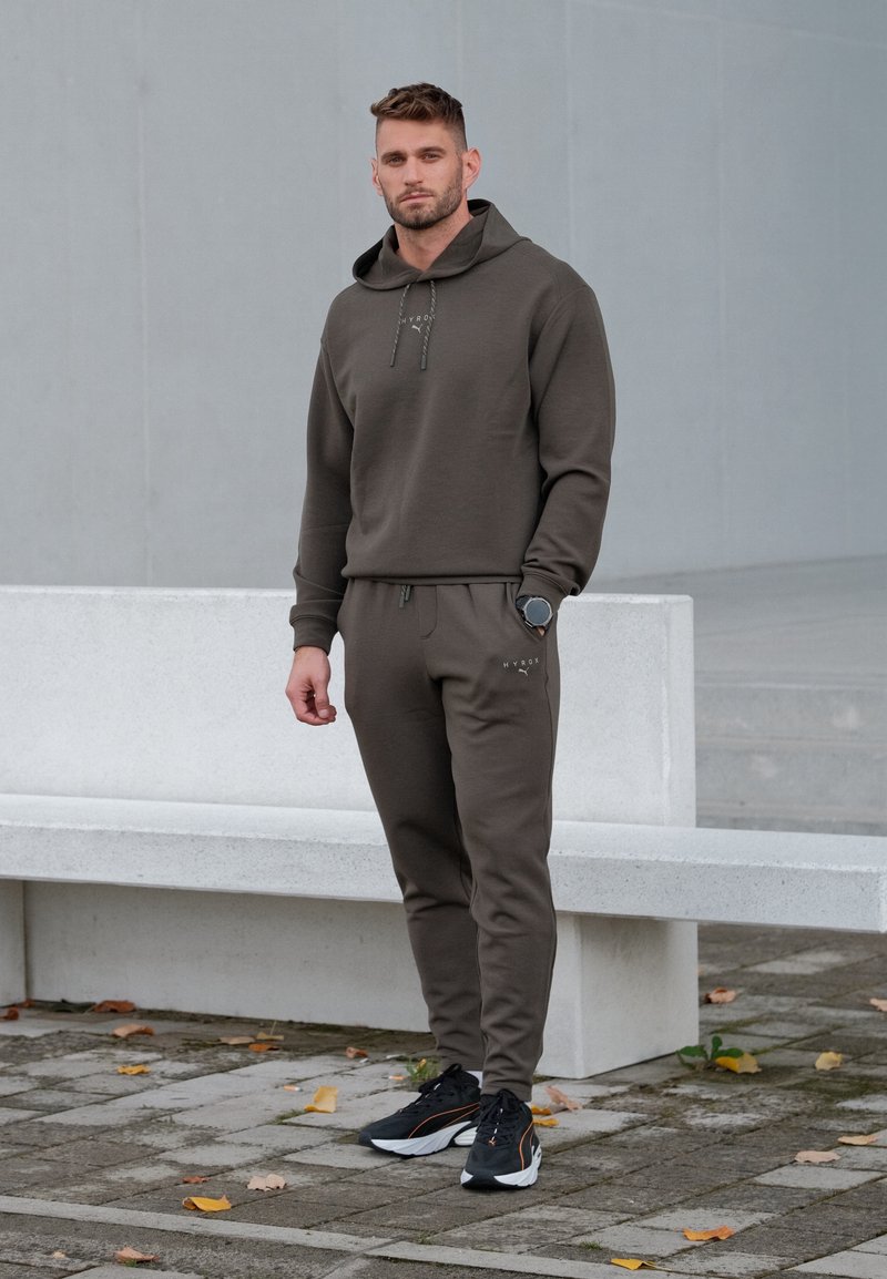 Sweat à capuche vert olive foncé avec cordons ajustables et joggeurs assortis. Tissu texturé, associé à des chaussures de sport noires avec des accents orange.