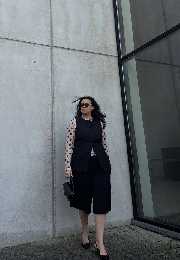 Žena so štýlovými čiernymi slnečnými okuliarmi, dlhým dierkovaným tričkom s bodkami, čiernou vestou, culottes nohavicami a na podpätkoch, s kabelkou, ktorá kráča okolo modernej betónovej a sklenej steny.