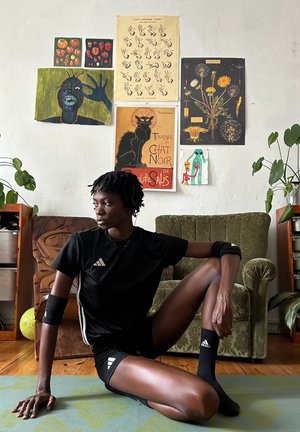 Pantalones cortos deportivos negros y camiseta con detalles blancos, acompañados de muñequeras y calcetines. El fondo presenta carteles coloridos y vegetación.