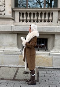 Femme en manteau marron doublé de fourrure, capuche en tricot crème et moufles, debout de profil sur un trottoir en ville devant un bâtiment en pierre avec une fenêtre grillagée.