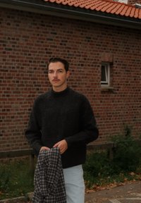 Schwarzer, strukturiert gestrickter Pullover mit hohem Kragen; hält ein grau-blau kariertes Hemd. Vor einer Ziegelmauer und Pflanzen.